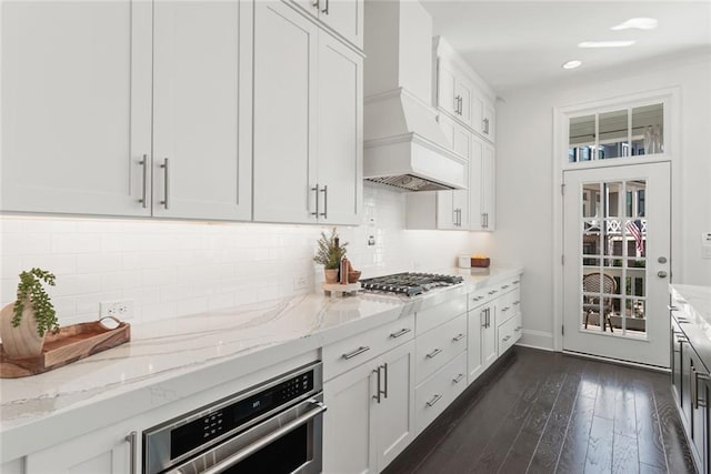 kitchen with light stone countertops, custom exhaust hood, dark wood-type flooring, appliances with stainless steel finishes, and tasteful backsplash