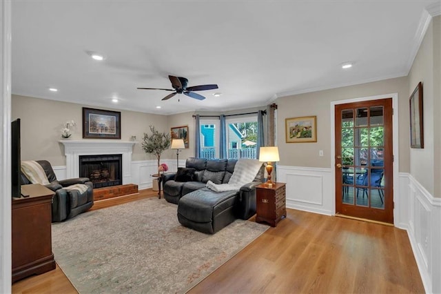 living room with a ceiling fan, crown molding, a wainscoted wall, a fireplace with raised hearth, and light wood-style flooring