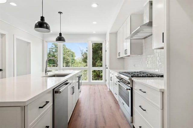 kitchen featuring sink, hanging light fixtures, stainless steel appliances, wall chimney range hood, and white cabinets