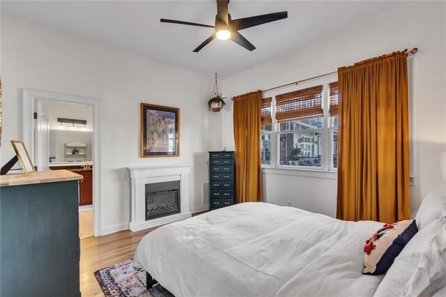 bedroom with connected bathroom, a ceiling fan, baseboards, light wood-style floors, and a glass covered fireplace