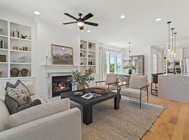 living room with a glass covered fireplace, light wood-style flooring, ceiling fan with notable chandelier, built in shelves, and recessed lighting