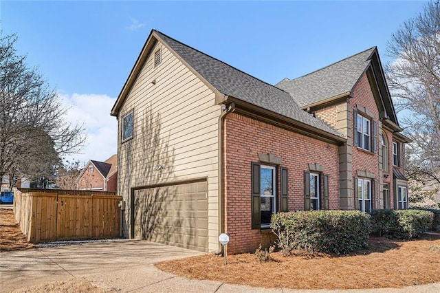 view of property exterior featuring driveway, fence, roof with shingles, a garage, and brick siding