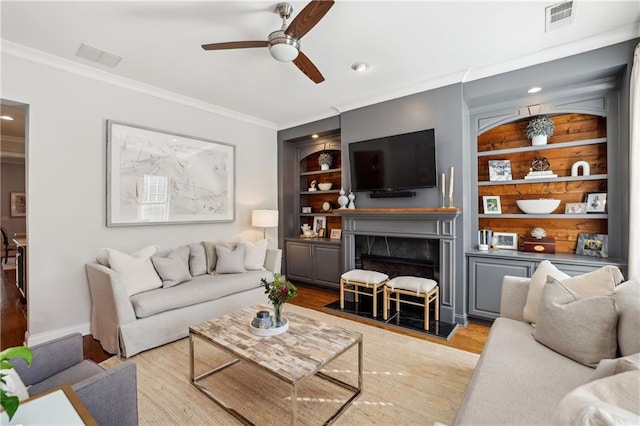 living room featuring a fireplace, visible vents, built in features, ornamental molding, and light wood-type flooring