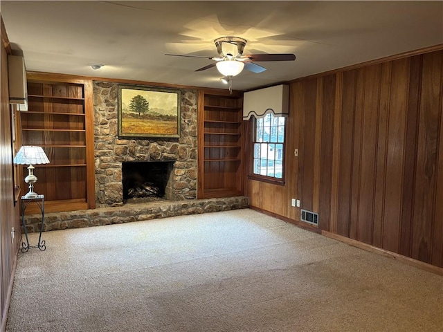 unfurnished living room with wood walls, visible vents, carpet floors, a ceiling fan, and a stone fireplace