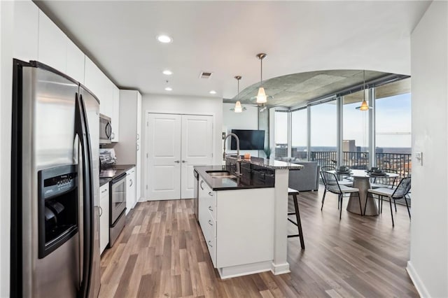 kitchen featuring white cabinetry, decorative light fixtures, a wall of windows, stainless steel appliances, and a kitchen island with sink