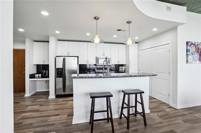 kitchen with white cabinetry, hanging light fixtures, dark stone countertops, appliances with stainless steel finishes, and an island with sink