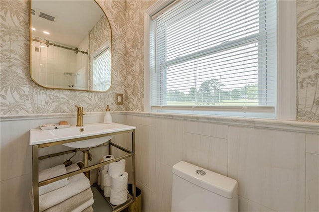 bathroom featuring wallpapered walls, a wainscoted wall, toilet, a shower, and a sink