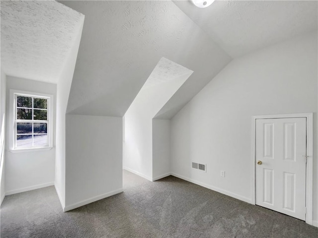 additional living space with visible vents, a textured ceiling, dark carpet, baseboards, and vaulted ceiling