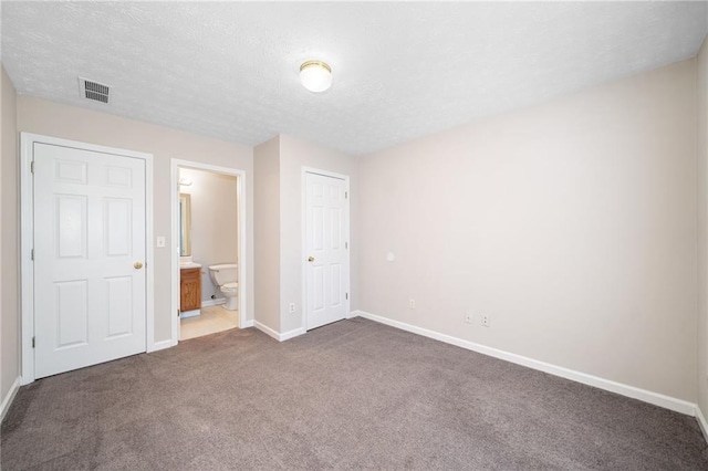 unfurnished bedroom with a textured ceiling, visible vents, baseboards, dark colored carpet, and ensuite bath