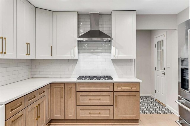 kitchen with wall chimney exhaust hood, stainless steel appliances, light countertops, white cabinetry, and backsplash