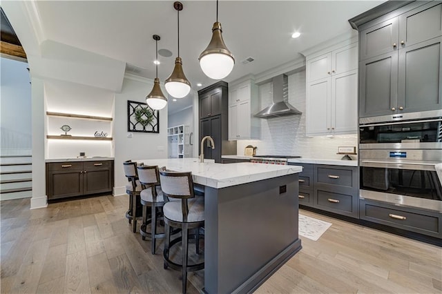 kitchen featuring a kitchen island with sink, a breakfast bar, wall chimney range hood, appliances with stainless steel finishes, and decorative light fixtures