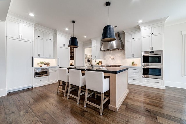 kitchen featuring dark countertops, an island with sink, wall chimney exhaust hood, stainless steel double oven, and white cabinetry