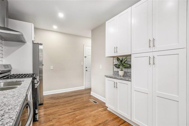 kitchen with white cabinetry, stainless steel appliances, light stone countertops, wall chimney exhaust hood, and light hardwood / wood-style flooring