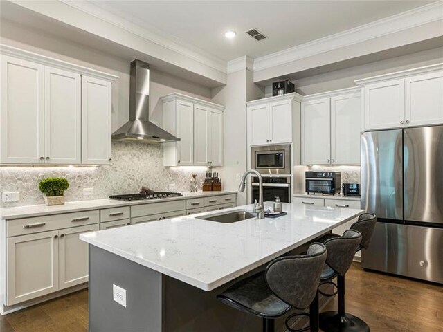 kitchen featuring appliances with stainless steel finishes, white cabinetry, wall chimney range hood, and a center island with sink