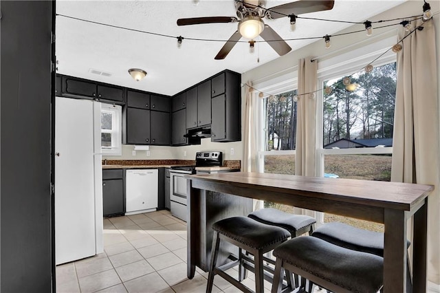 kitchen featuring light tile patterned flooring, under cabinet range hood, dark cabinets, white appliances, and visible vents