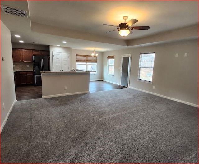 unfurnished living room with ceiling fan with notable chandelier, dark carpet, and a tray ceiling