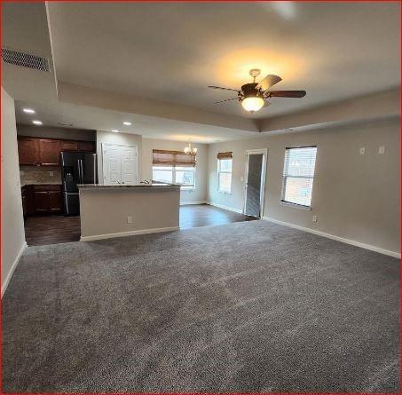 unfurnished living room featuring a raised ceiling, ceiling fan with notable chandelier, and dark colored carpet