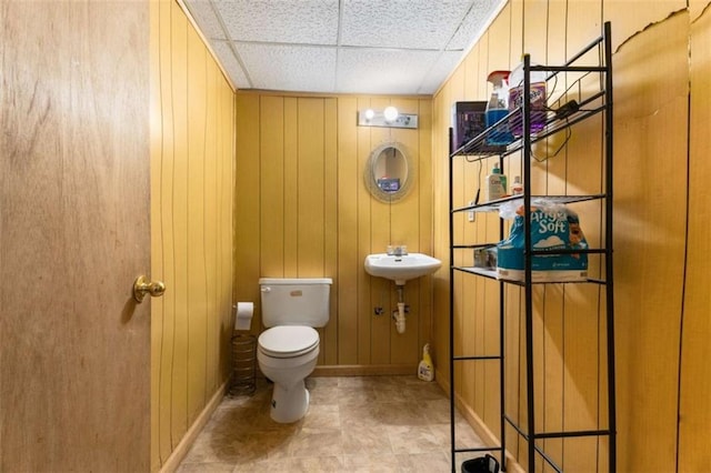 half bath with baseboards, a sink, a paneled ceiling, wood walls, and toilet