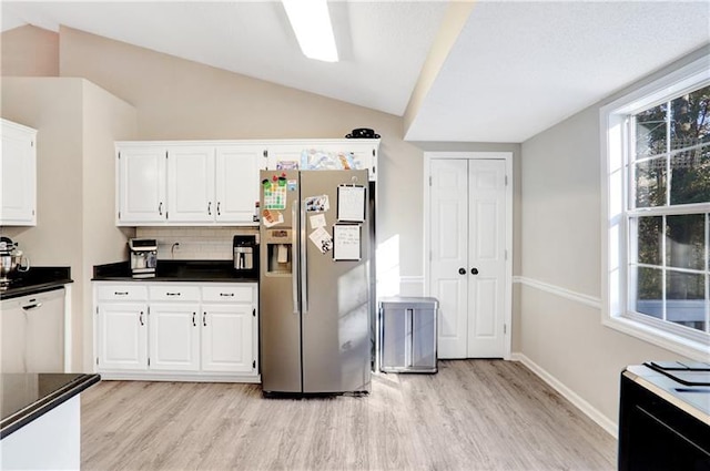 kitchen with stainless steel fridge, dark countertops, lofted ceiling, white cabinetry, and light wood-style floors