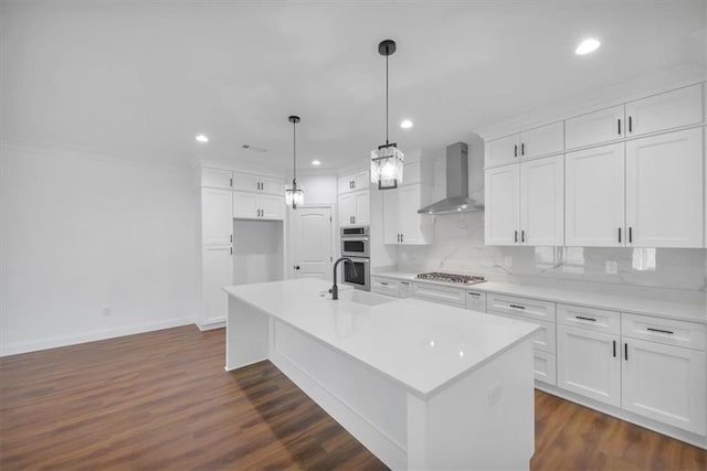 kitchen with wall chimney range hood, white cabinetry, a center island with sink, and appliances with stainless steel finishes