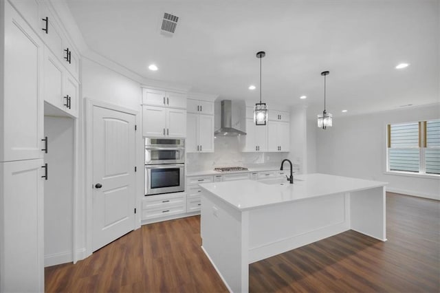 kitchen with pendant lighting, appliances with stainless steel finishes, white cabinetry, wall chimney range hood, and a center island with sink