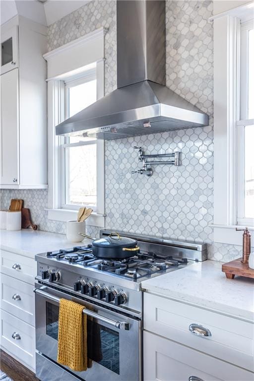 kitchen with stainless steel range, glass insert cabinets, wall chimney exhaust hood, white cabinets, and decorative backsplash