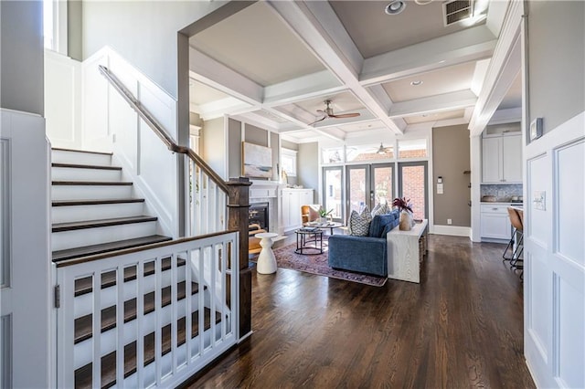 living area with stairway, a ceiling fan, coffered ceiling, beam ceiling, and a glass covered fireplace