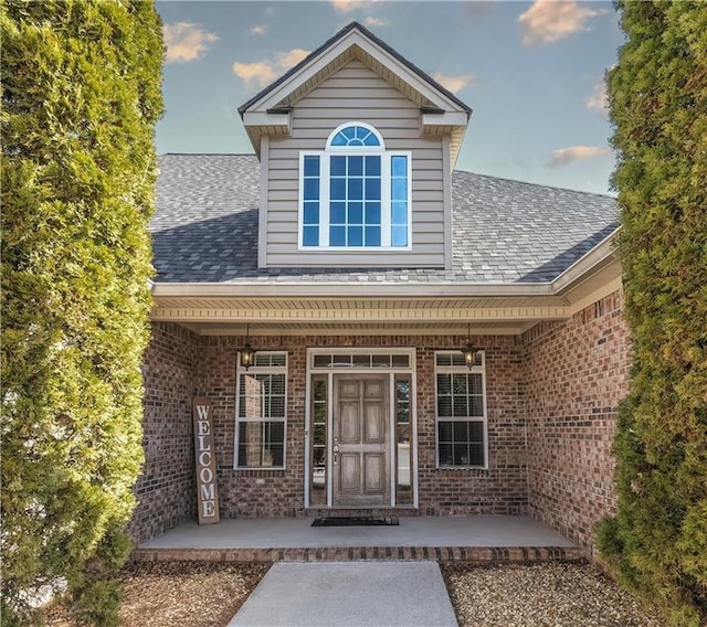 entrance to property featuring covered porch, brick siding, and a shingled roof