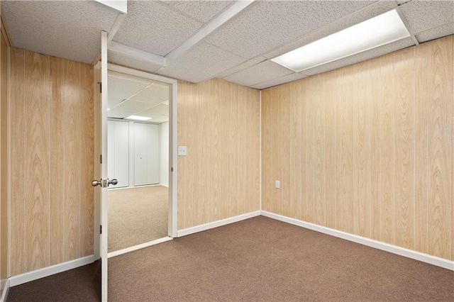 basement featuring dark colored carpet, a paneled ceiling, and wood walls