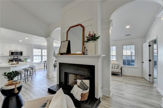 living room featuring high vaulted ceiling, light wood-type flooring, a brick fireplace, and ornate columns