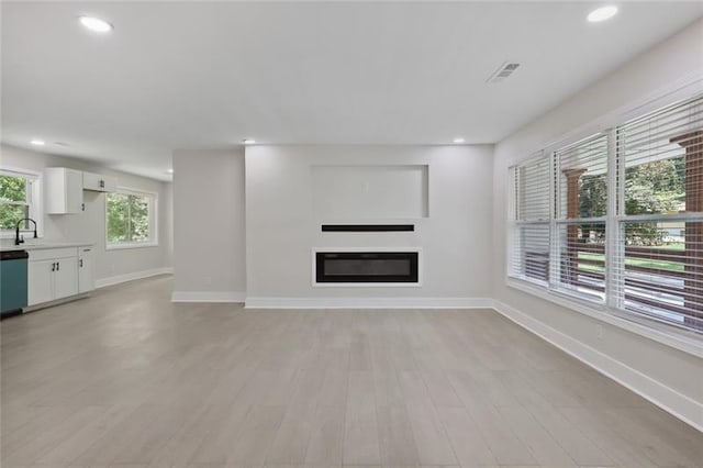 unfurnished living room with light wood-type flooring, a sink, baseboards, a glass covered fireplace, and recessed lighting
