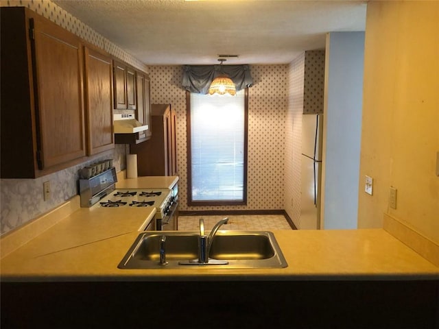 kitchen with gas stove, under cabinet range hood, freestanding refrigerator, wallpapered walls, and a sink