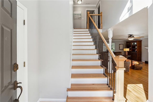 stairway featuring wood finished floors, a ceiling fan, baseboards, decorative columns, and crown molding