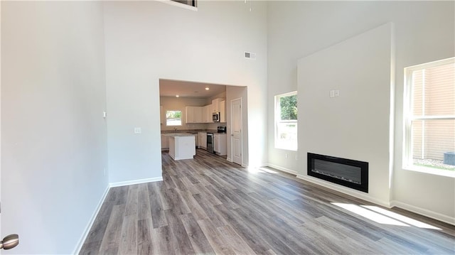 unfurnished living room featuring visible vents, light wood-style flooring, a high ceiling, a glass covered fireplace, and baseboards