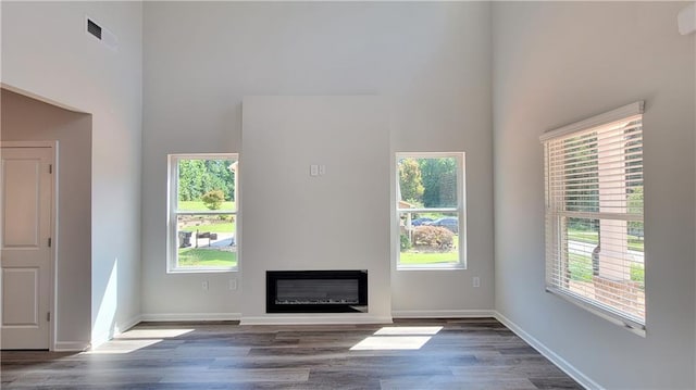 unfurnished living room with baseboards, visible vents, a glass covered fireplace, a towering ceiling, and wood finished floors