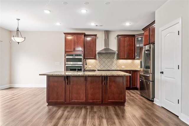 kitchen with wall chimney range hood, stainless steel appliances, wood finished floors, light stone counters, and backsplash