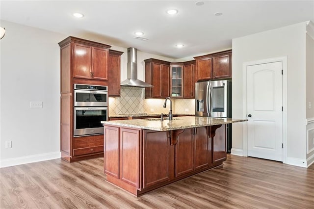 kitchen with wall chimney exhaust hood, a sink, stainless steel appliances, wood finished floors, and light stone countertops