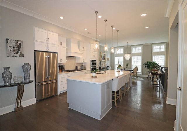 kitchen with hanging light fixtures, premium range hood, white cabinetry, high end refrigerator, and a center island with sink