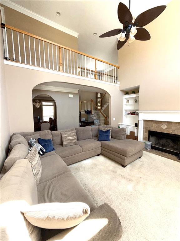 living area with arched walkways, a tiled fireplace, ornamental molding, a ceiling fan, and stairs