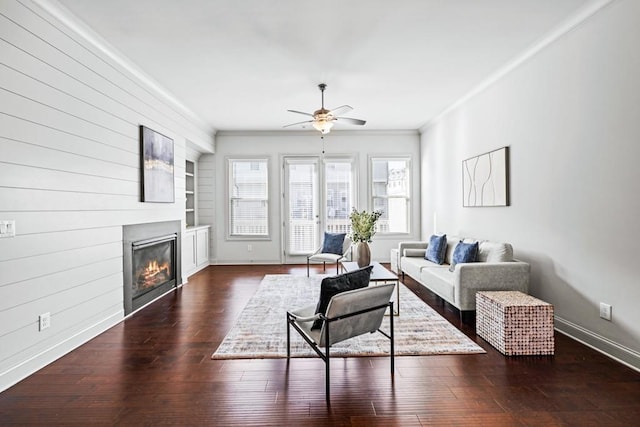 living area featuring built in shelves, dark wood-style flooring, ornamental molding, a glass covered fireplace, and baseboards