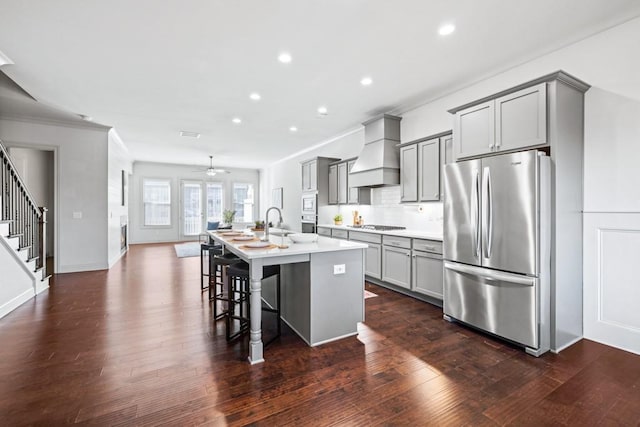 kitchen with a center island with sink, custom exhaust hood, stainless steel appliances, gray cabinetry, and a kitchen bar