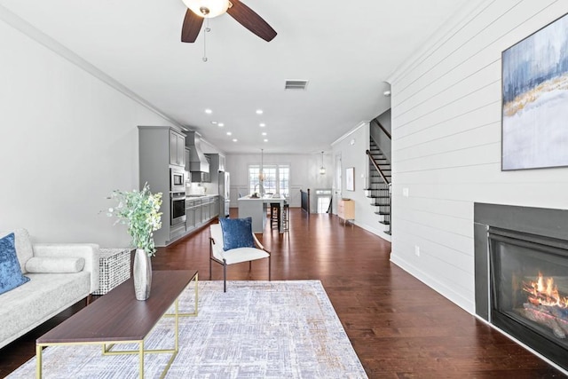 living room featuring dark wood-style floors, visible vents, a ceiling fan, a glass covered fireplace, and stairs