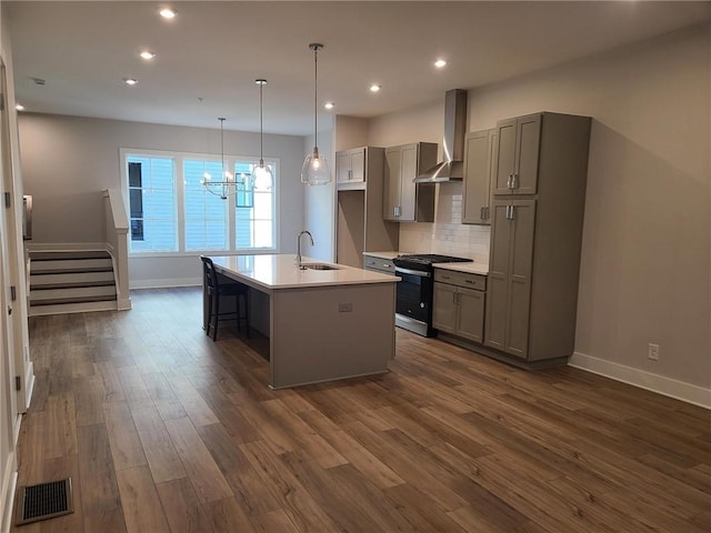 kitchen featuring dark hardwood / wood-style flooring, wall chimney exhaust hood, stainless steel gas range, decorative light fixtures, and an island with sink