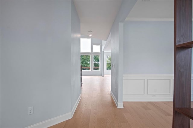 hall with light wood finished floors, baseboards, a wainscoted wall, ornamental molding, and a decorative wall