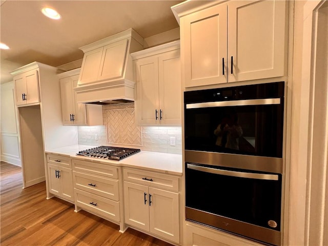 kitchen featuring backsplash, white cabinetry, stainless steel appliances, custom range hood, and hardwood / wood-style flooring