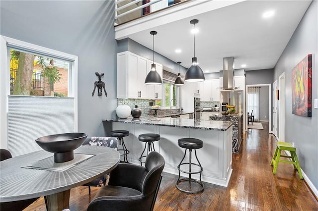 kitchen featuring dark stone counters, wall chimney exhaust hood, tasteful backsplash, decorative light fixtures, and white cabinetry