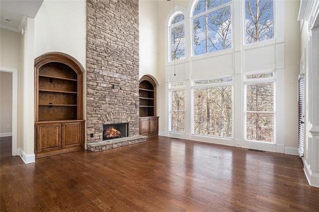 unfurnished living room with a towering ceiling, crown molding, dark wood-type flooring, a stone fireplace, and built in shelves