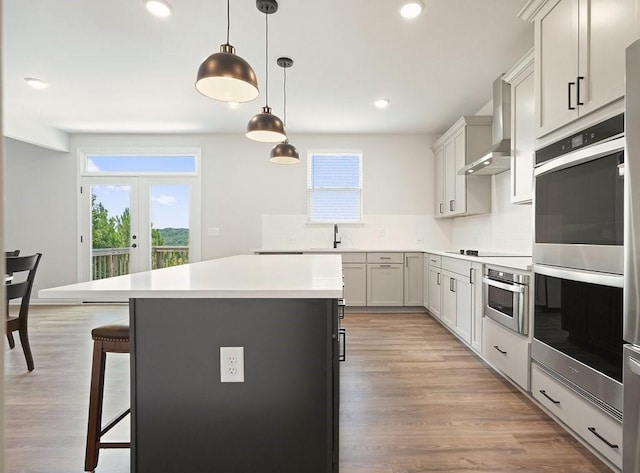 kitchen featuring pendant lighting, stainless steel double oven, a kitchen island, light hardwood / wood-style flooring, and wall chimney range hood