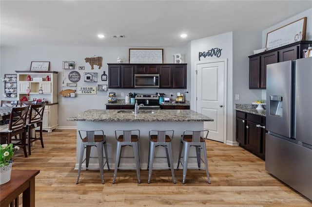 kitchen with dark brown cabinets, light stone counters, a kitchen island with sink, and appliances with stainless steel finishes