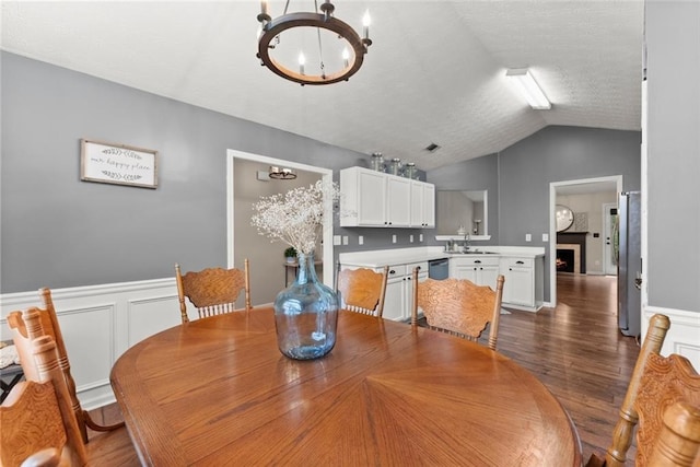 dining room with a warm lit fireplace, wainscoting, dark wood-type flooring, vaulted ceiling, and a chandelier